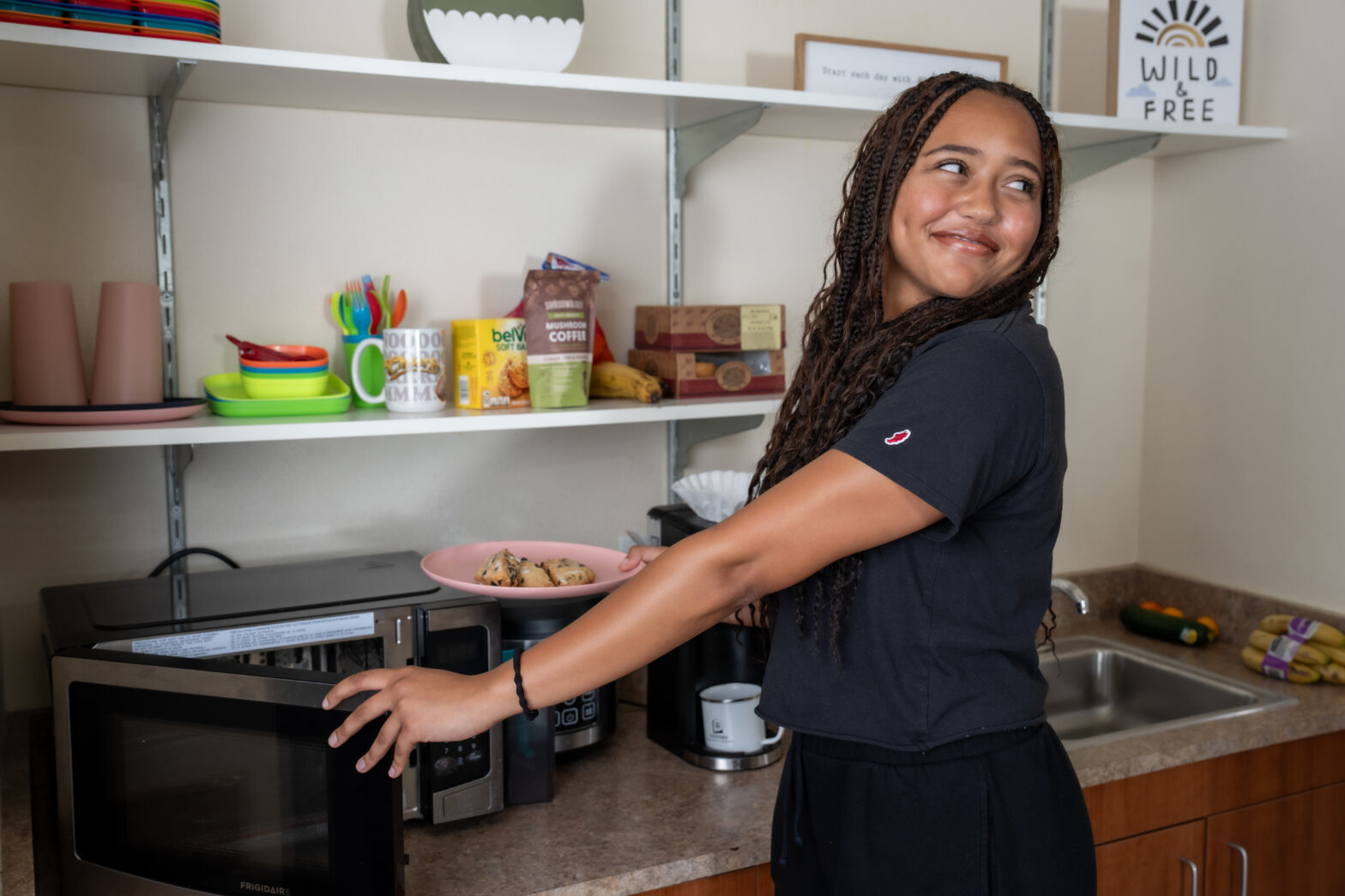 A young woman at a microwave with food, smiling.
