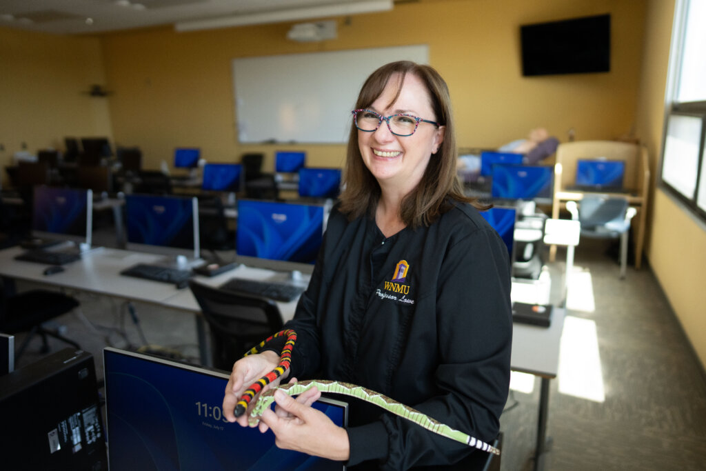 A female nurse with dark hair and glasses holds two separate types of snake models - one with red and black stripes, one with white and black stripes.
