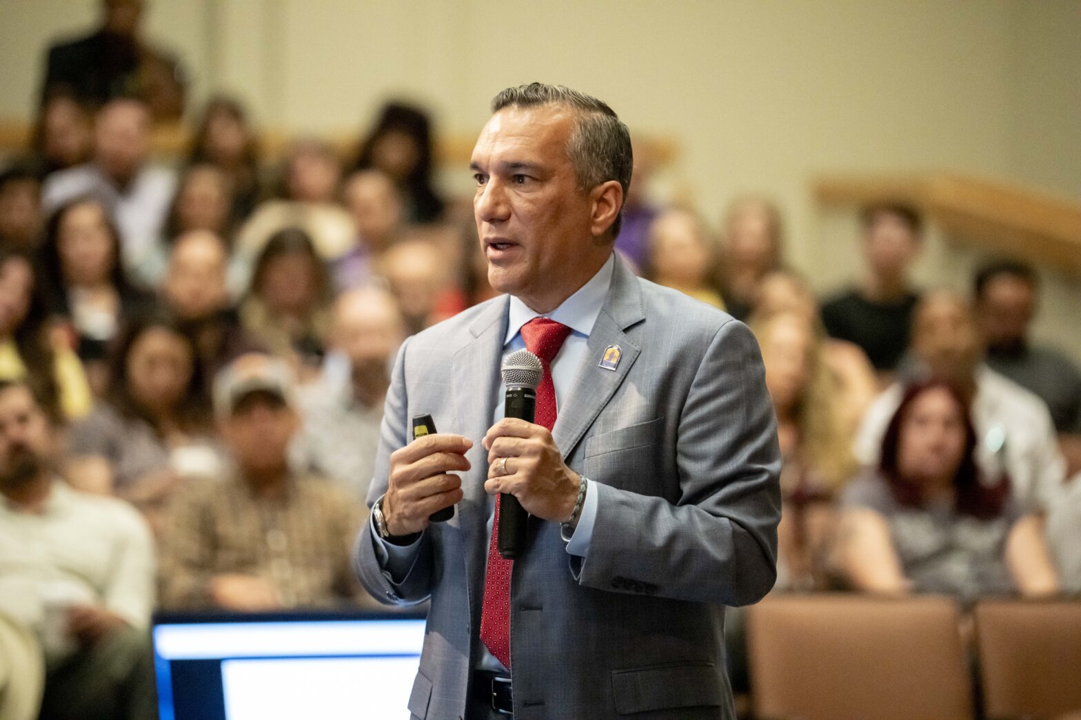 A man in a grey suit and red tie holding a microphone and addressing a crowd