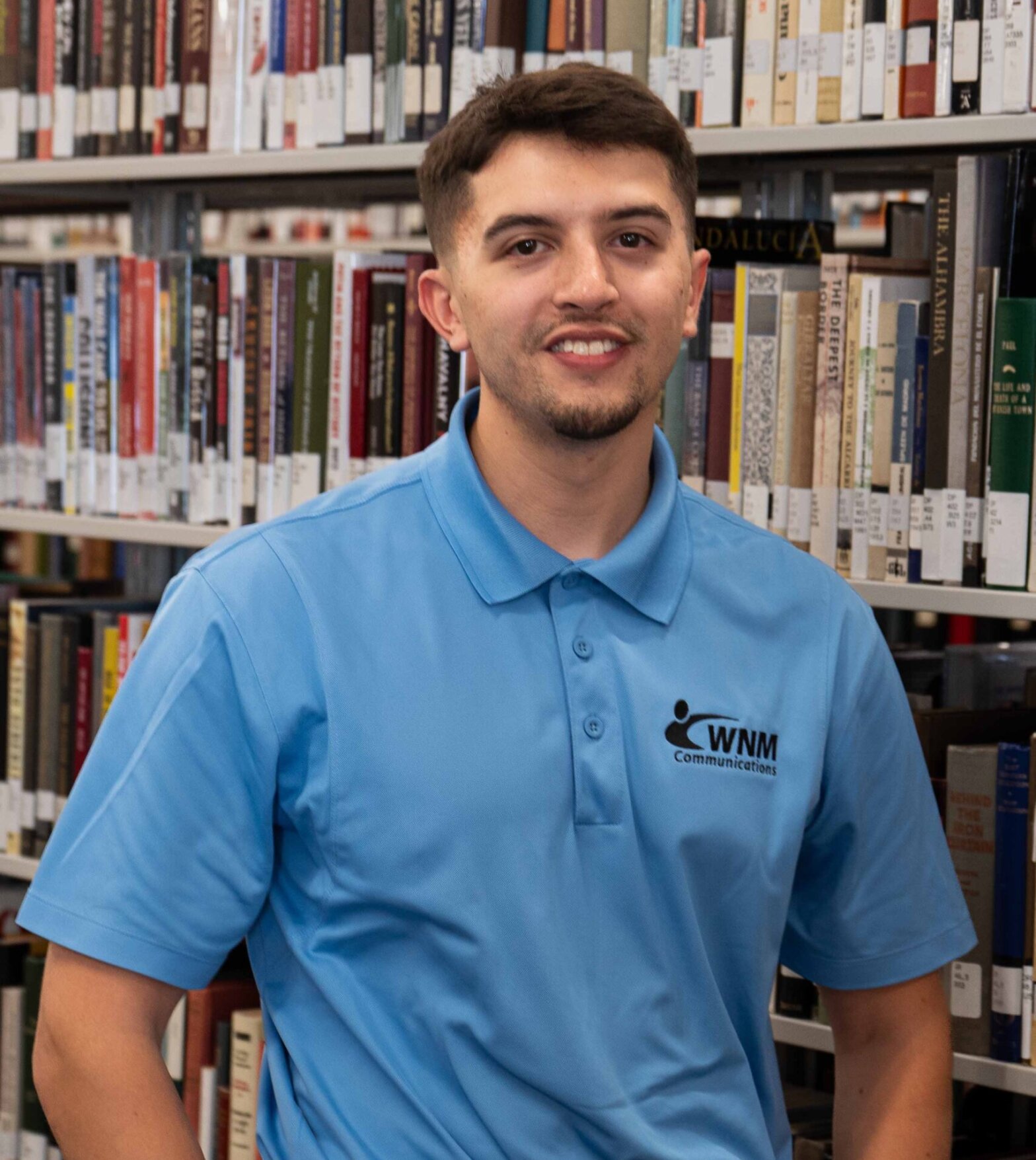 A young man with black hair in a blue shirt, standing against stacks of books.