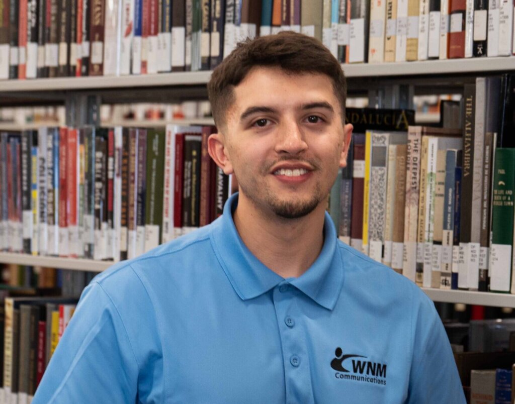 A young man with black hair in a blue shirt, standing against stacks of books.