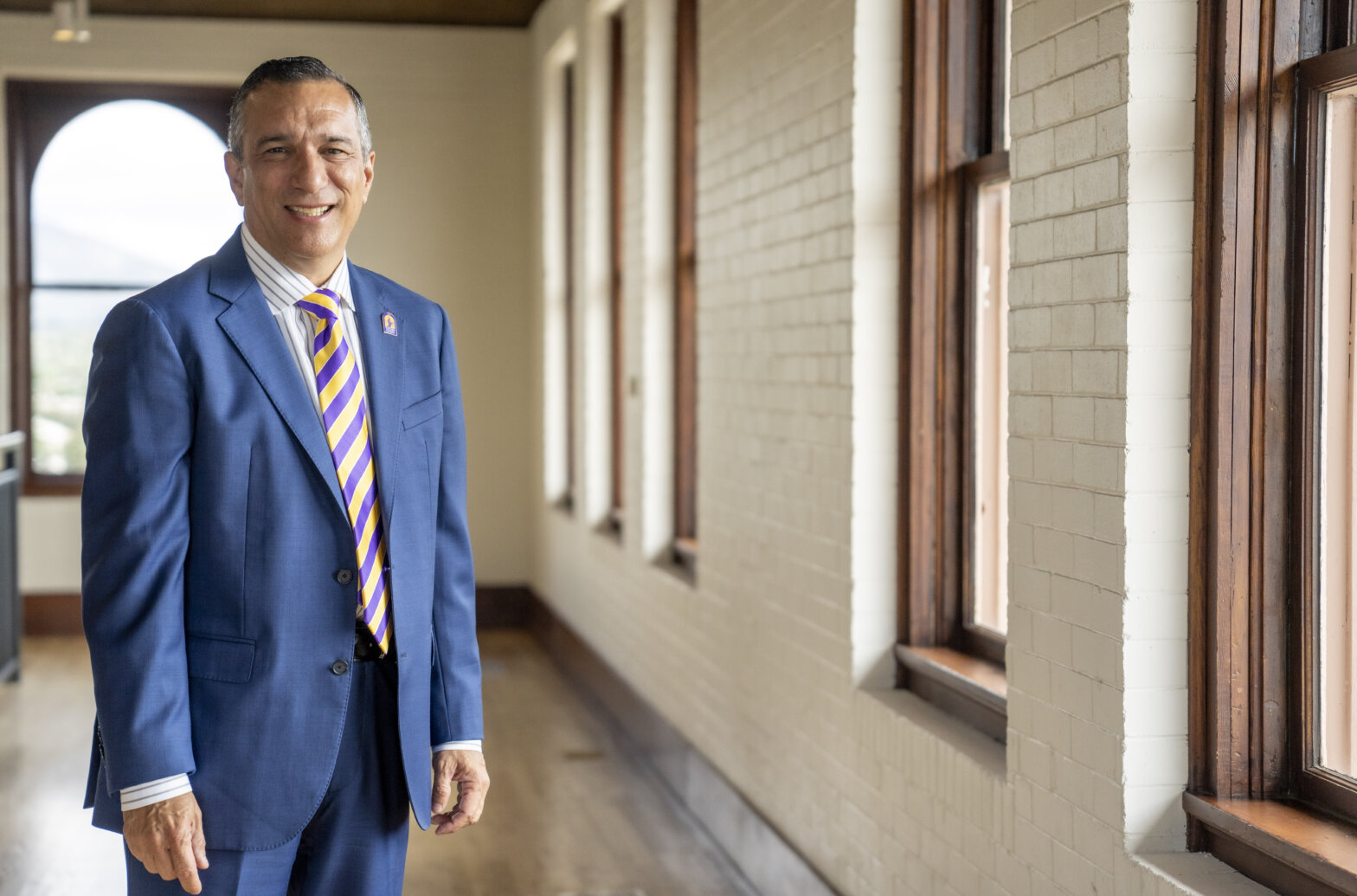 A man in a suit standing in a hallway next to large windows.