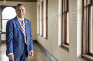 A man in a suit standing in a hallway next to large windows.